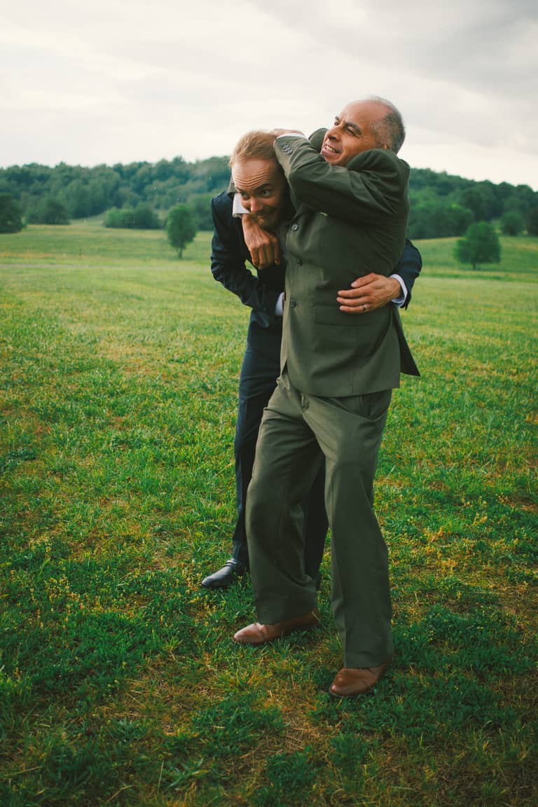 Two men in suits playfully wrestle in a grassy field with trees in the background