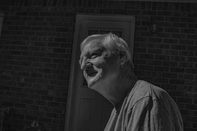 Black and white portrait of an older man smiling outdoors against a brick wall