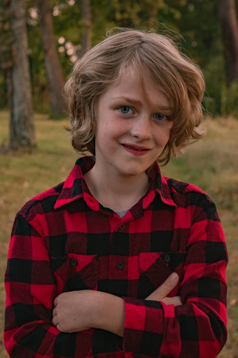 Young boy with blue eyes and freckles smiles in a red plaid shirt outdoors