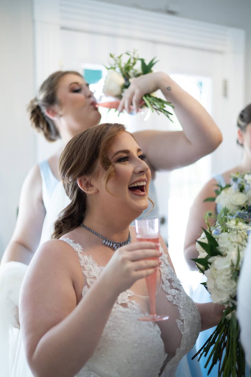 Bride and bridesmaids laugh while holding champagne and bouquets at a wedding