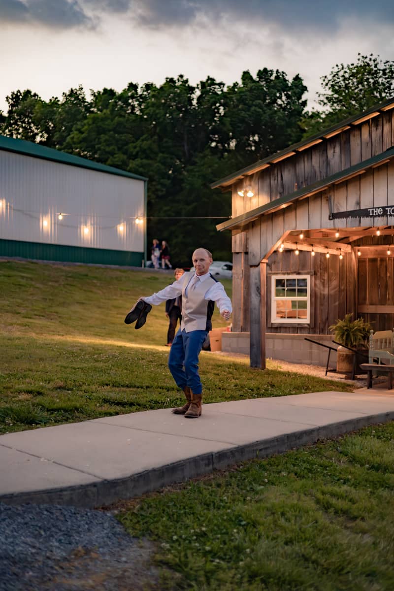 Groom dances joyfully outside a rustic barn at dusk, holding his shoes