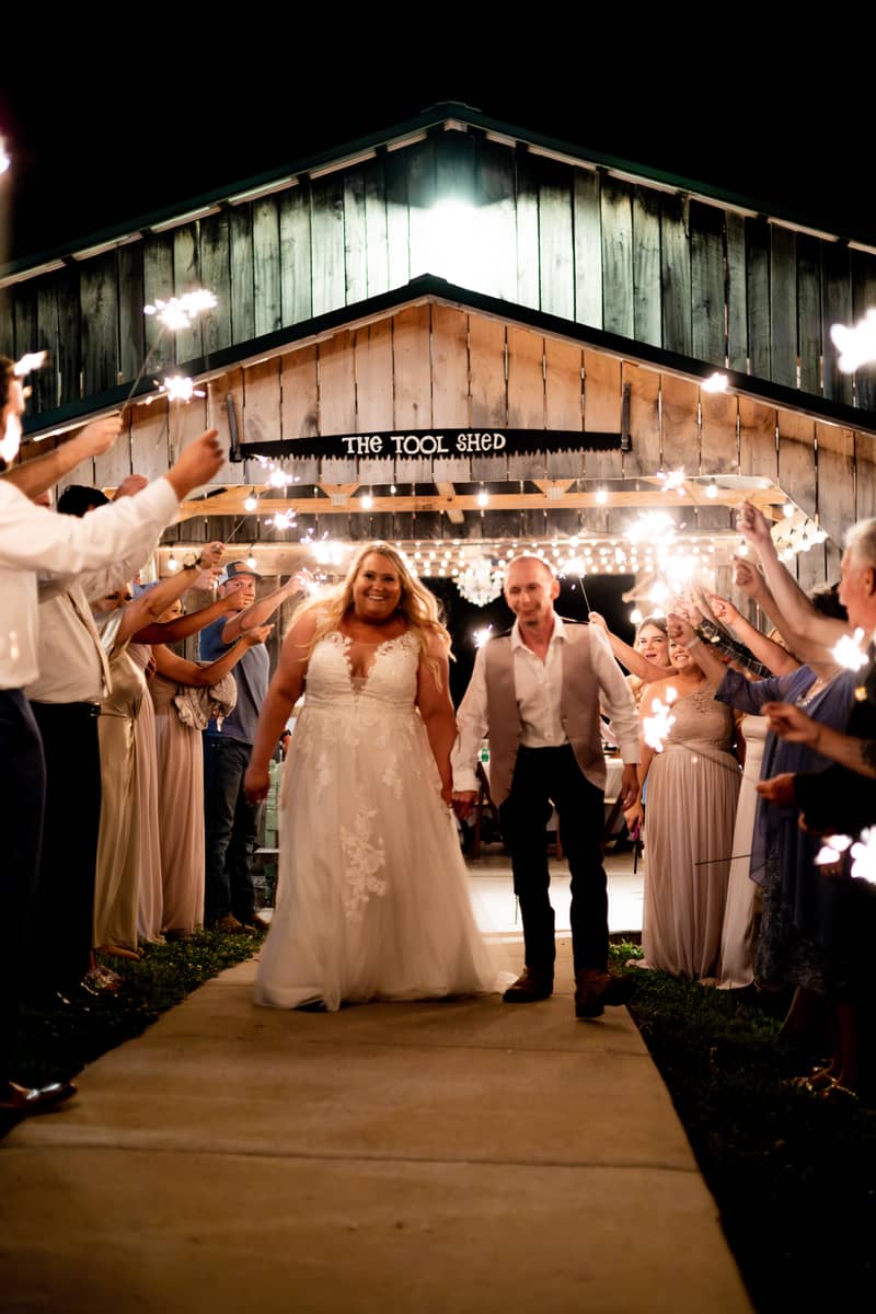 Newlyweds walk through a sparkler exit at night in front of a rustic venue