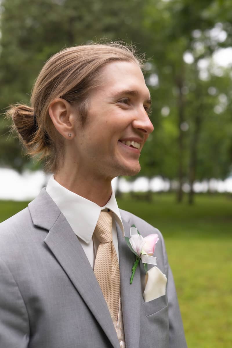 Groom smiles outdoors in a grey suit with a boutonniere during his wedding day