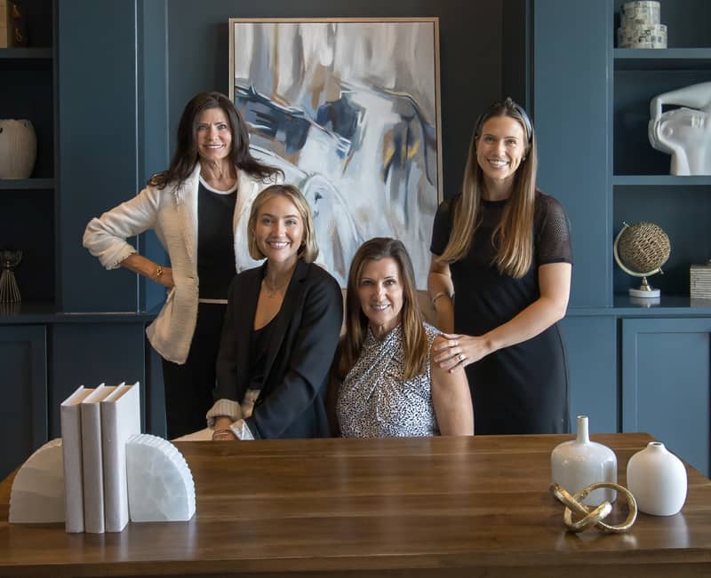 Four professional women smile confidently in a modern office setting with abstract art