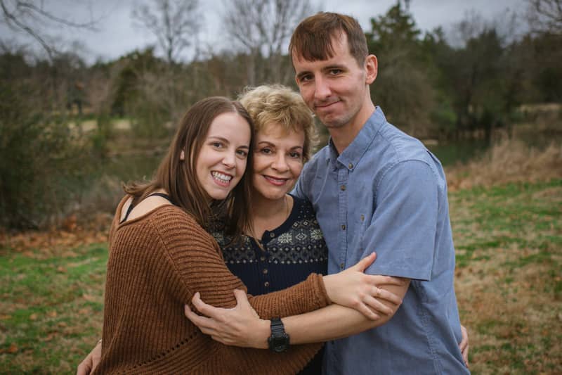 Three smiling family members embrace outdoors near a body of water