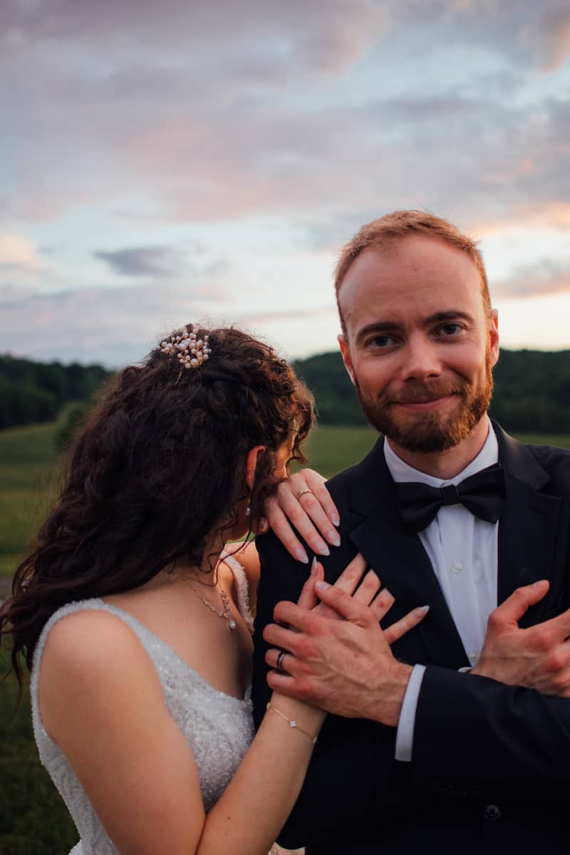Groom smiles at camera while bride rests head on his shoulder during a romantic wedding portrait