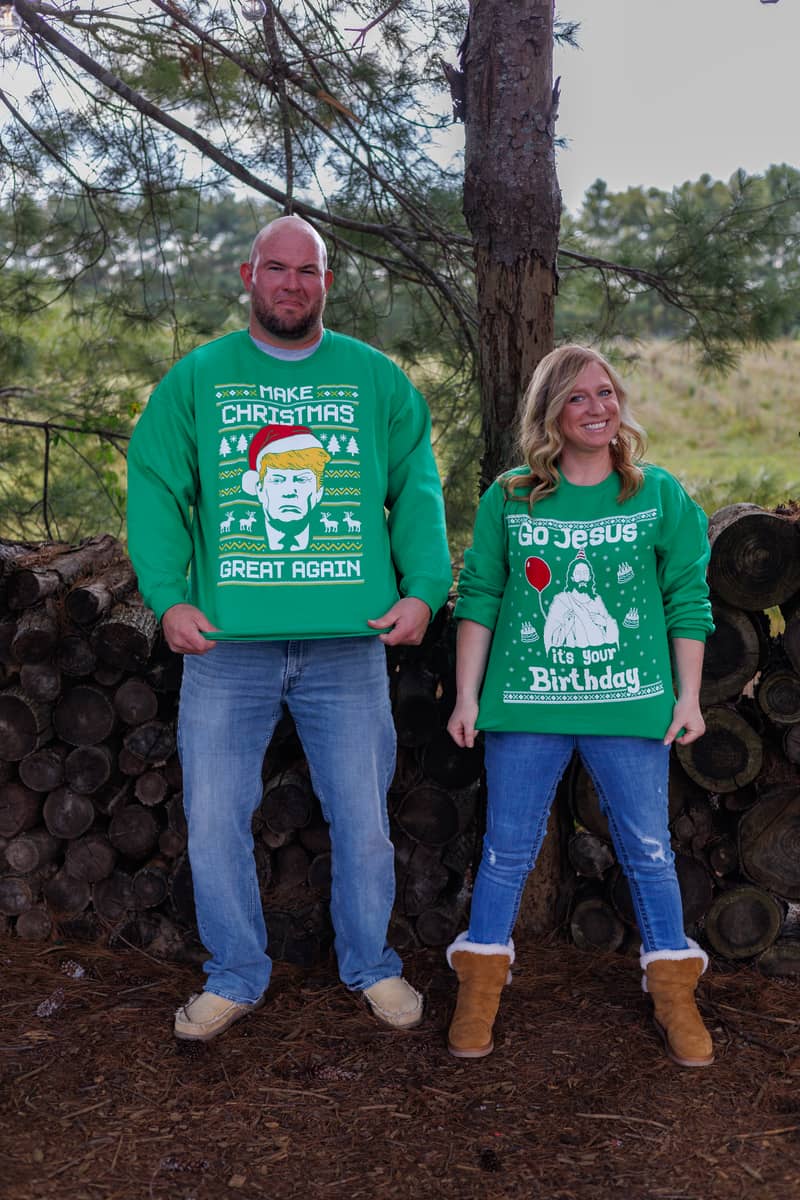 Couple in festive green sweaters poses outdoors with a woodpile background