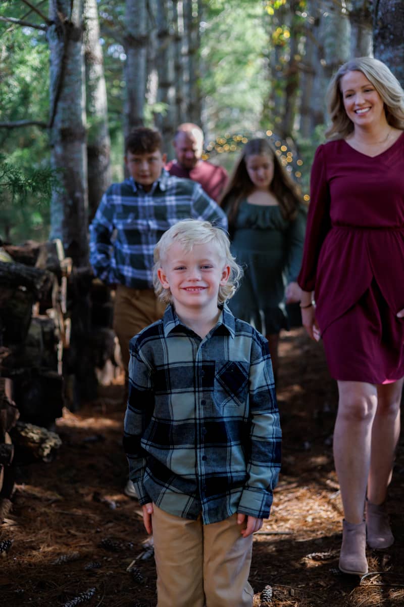 A smiling boy walks toward the camera in a forest with his family behind him